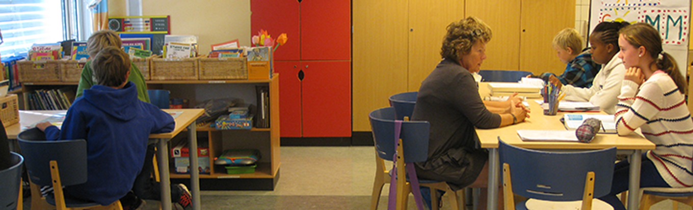 Bilde av klasserommet ved Ullevål. Det er flere elever i klasserommet, og en lærer sitter ved et av bordene og underviser en elev.